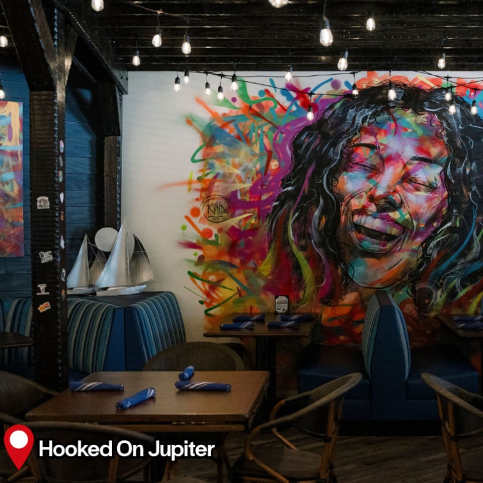 HOOKED restaurant interior with colorful mural, string lights, wooden tables, and blue striped seating.
