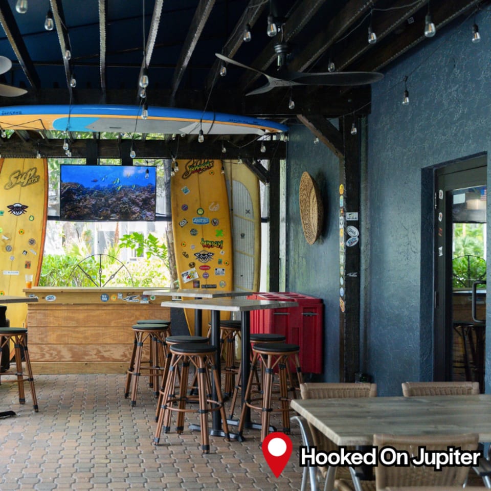 HOOKED's casual outdoor bar area with surfboards, high-top tables, stools, and string lights.