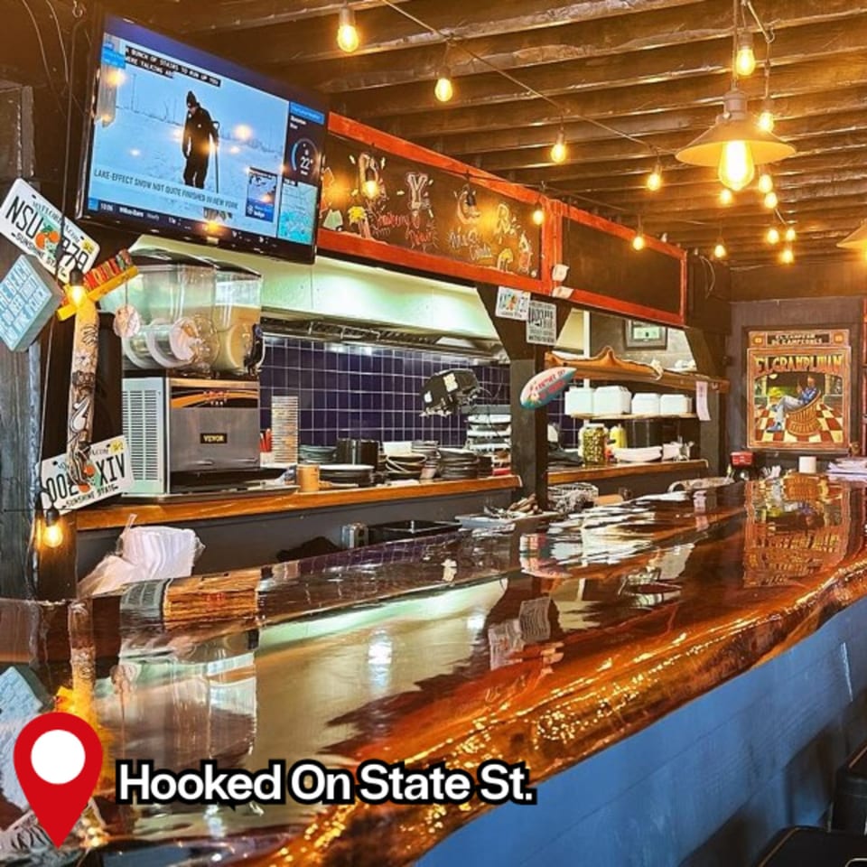 A long, glossy wooden bar with a television, kitchen area, and string lights in Hooked restaurant.